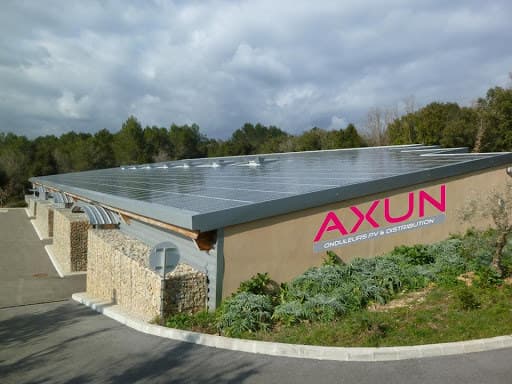 Bâtiment d’AXUN vu de trois quarts : longue toiture inclinée entièrement couverte de panneaux solaires, enseigne « AXUN — onduleurs PV & distribution » sur la façade, pinède à l’arrière-plan.