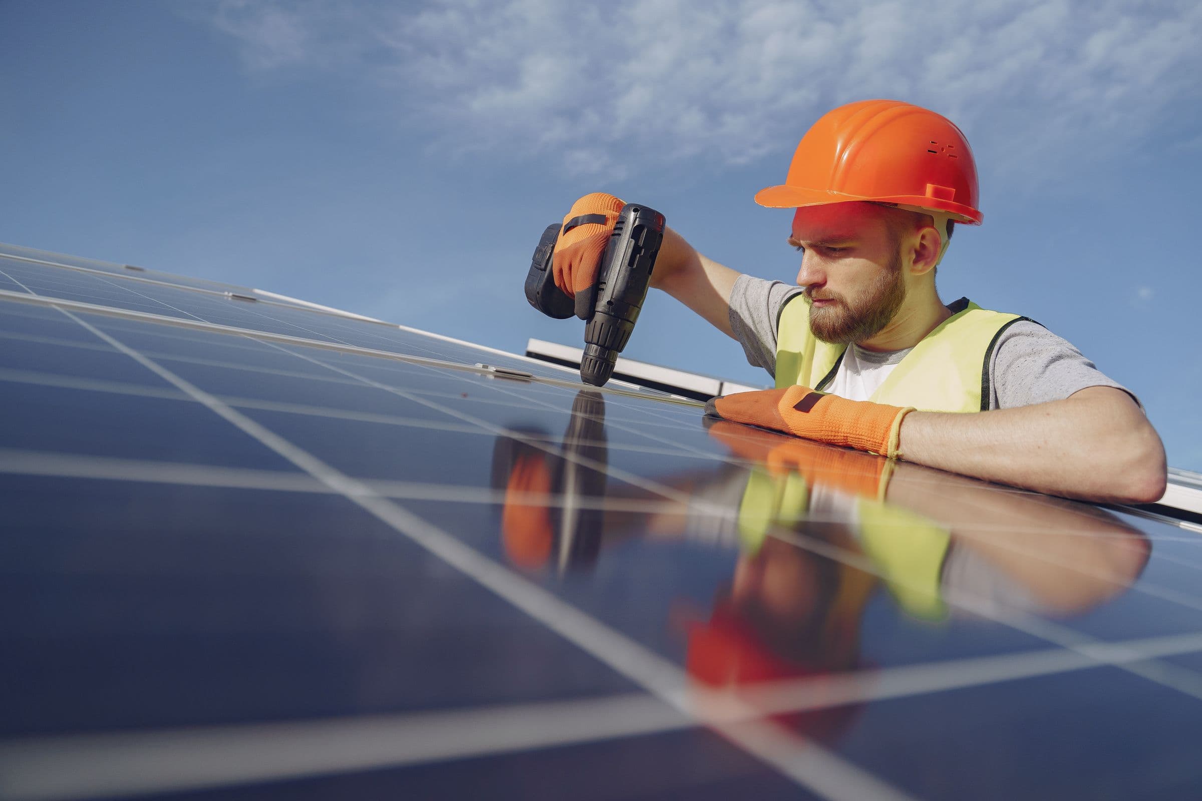 Technicien en casque et gants de protection fixant un panneau solaire à la visseuse, sous un ciel bleu.