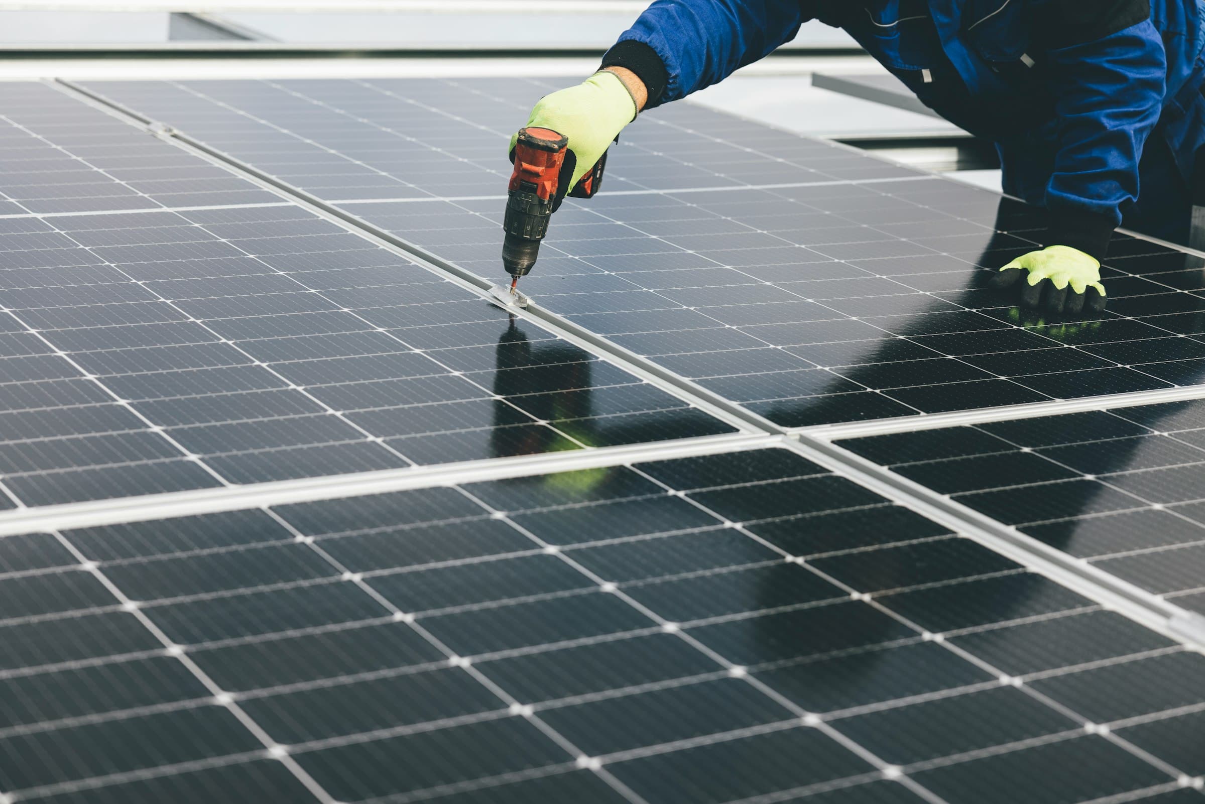 Gros plan d’un technicien qui visse un rail de fixation de panneau solaire avec une visseuse électrique, gants de protection.