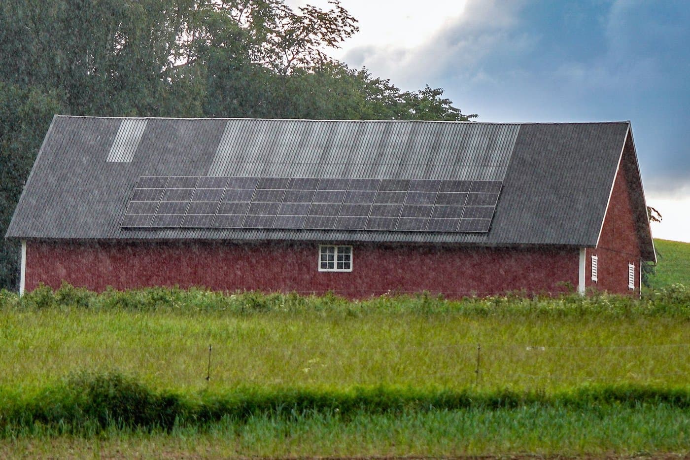 Grange agricole isolée en bois rouge sous l’averse, longue rangée de panneaux solaires sur le pan du toit.