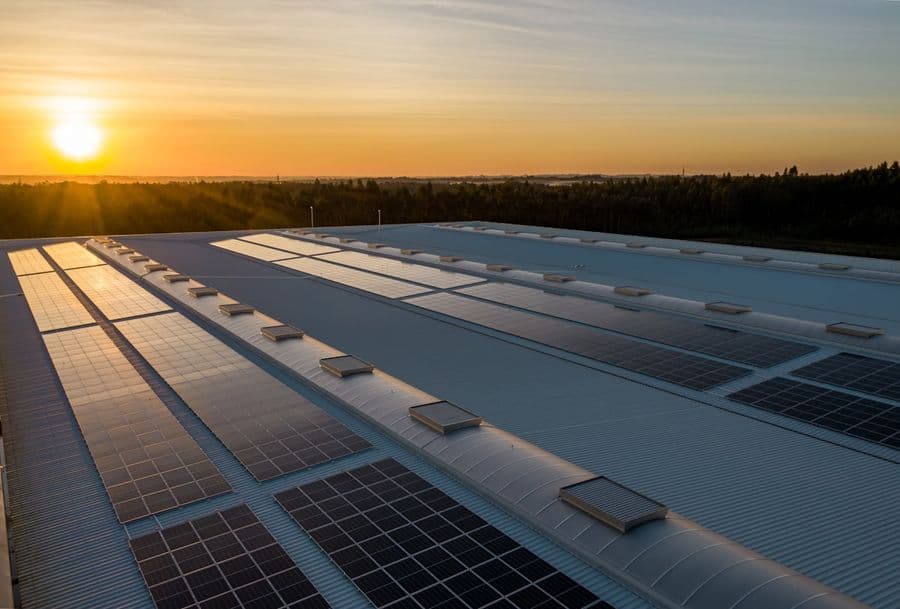 Vue aérienne d'une toiture industrielle courbe équipée de plusieurs rangées de panneaux photovoltaïques, au lever du soleil.
